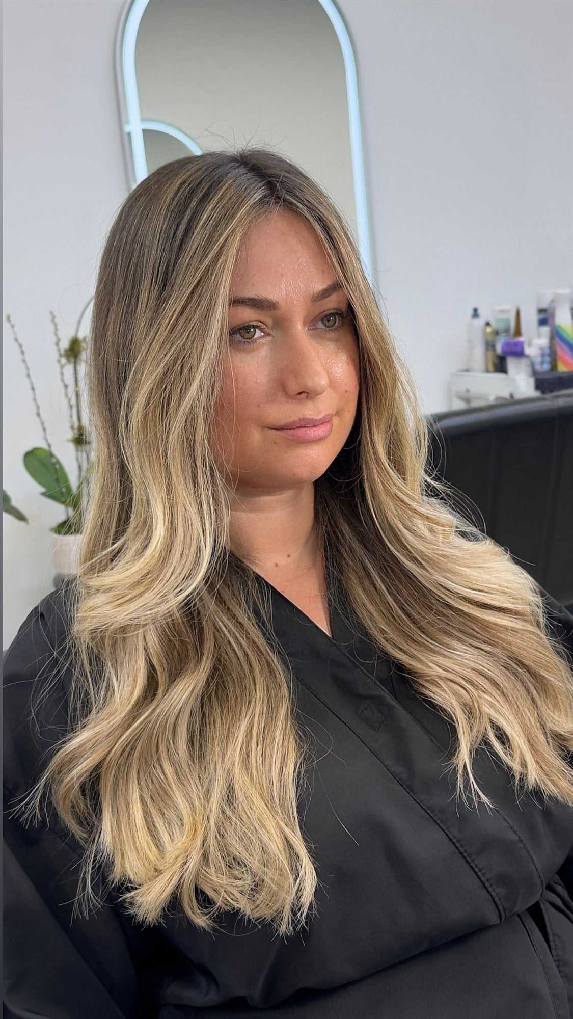 Woman with long, wavy blonde hair in salon, wearing black cape. Modern decor in background.