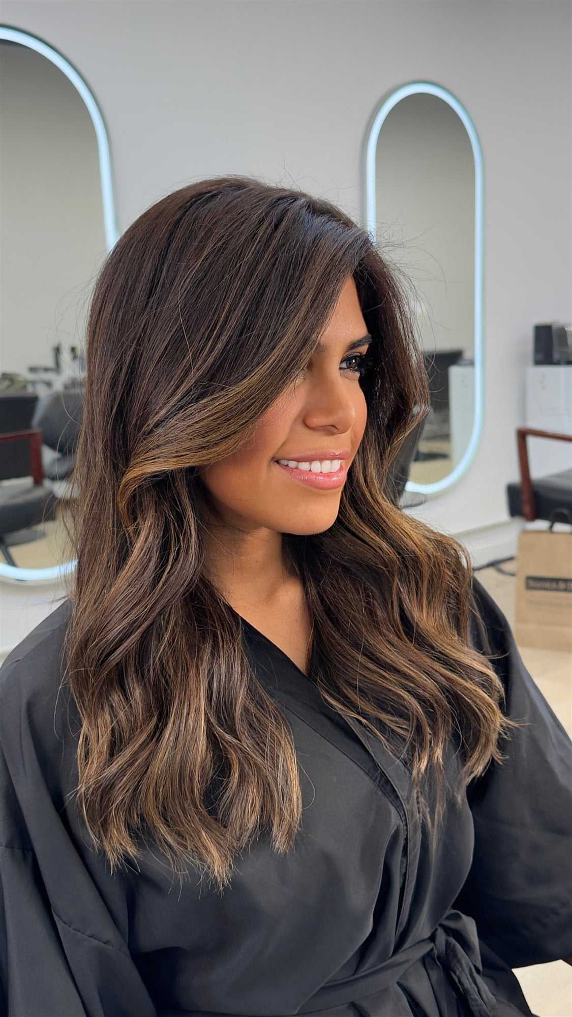 Smiling person with long brown hair in a salon, wearing a black robe.