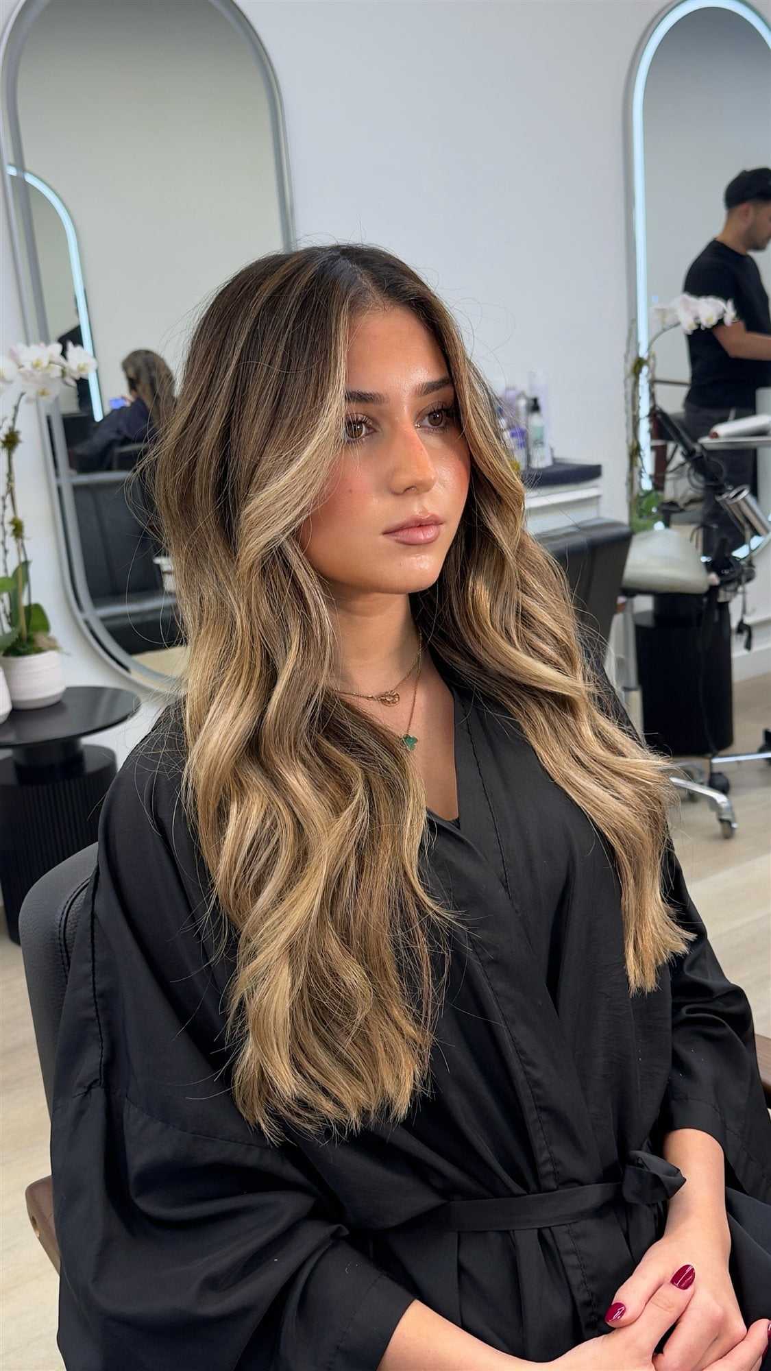 Woman with long, wavy hair sits in a salon, mirrors and orchids in the background.