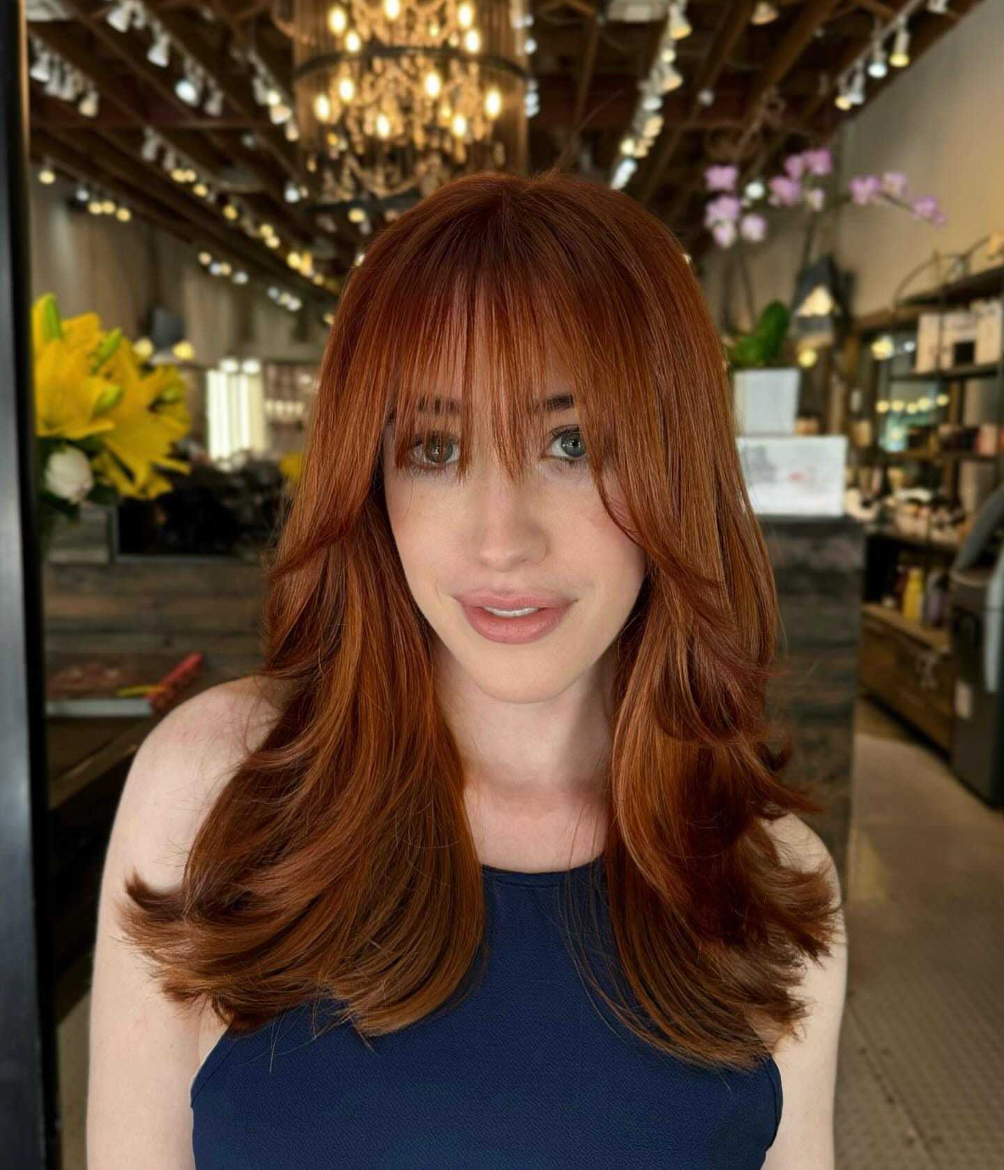 Red-haired woman with bangs in a chic salon, elegant lighting and floral decor in background.